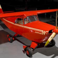 Diagonal front view of red monoplane with yellow accent colors and one engine. Fixed landing gear is used to make the aircraft roadable.