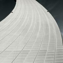 Partial view of the air traffic control tower at Abu Dhabi International Airport. The base has a curved, white tile pattern with the top leading to a metal control tower base.