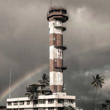 Partial view of an air traffic control tower at Ford Island Airport in Hawaii. The base features an alternating white and brown-colored concrete with three open balconies. Above the base, a single story exists for monitoring aircrafts and a balcony is found throughout the top of the tower.