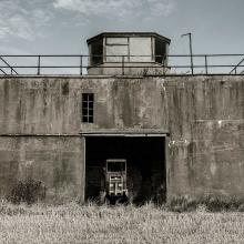 Partial view of the air traffic control tower at East Fortune Field in Scotland. The tower is three stories and features a concrete base with an opening in the center of the first floor. The third floor features at least four different observation angles.