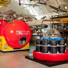 A large red and yellow balloon gondola with a rectangular shape is next to a red and black gondula with a smaller, red circular base and black supports on top of the base. They are both placed in the Museum's Steven F. Udvar-Hazy Center.