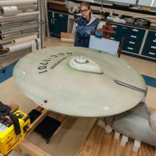 Top view of the starship Enterprise, a gray-colored, dish-shaped starship studio model, with a Smithsonian staff member standing near the studio model. The studio model is undergoing an X-ray scan.