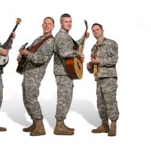 A group of four men in Army uniform pose informally with their instruments. The person on the far left is playing a banjo while everyone else seems to be playing guitar.