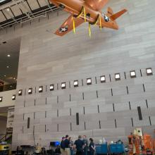 Several Museum employees stand under an orange monoplane hanging on the ceiling of the Museum's Boeing Milestones of Flight Hall. Nearby, an orange machine used to lift employees toward the aircraft is stationary.