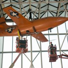 Two lifts are employed so that Museum employees can securely move the Bell X-1, an orange monoplane, from the ceiling to the floor of the Museum's Boeing Milestones of Flight Hall.