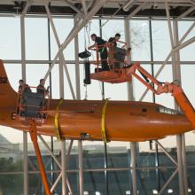 Using two lift machines, Museum employees work to secure the Bell X-1, an orange military monoplane, from the ceiling of the Boeing Milestones of Flight Hall onto the floor.