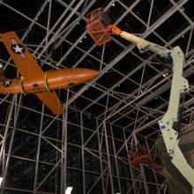 Using a singular lift machine, Museum employees work overnight to lower the Bell X-1, an orange military monoplane, from the ceiling to the floor of the Boeing Milestones of Flight Hall.