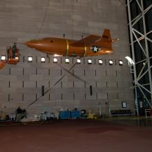 Museum employees in the middle of lowering the Bell X-1, an orange military monoplane, from the ceiling to the floor of the Boeing Milestones of Flight Hall. The aircraft is partially lowered.