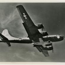 Bottom view of the Bell X-1, a military aircraft, being lifted under a B-29, a larger military aircraft. The B-29 is in flight.