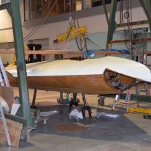 Diagonal side view of part of a white and wooden-colored glider aircraft, which is placed in between two metal frames. A section of clear plastic is removed from the front of the aircraft.