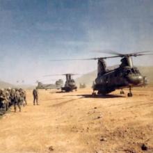 A group of soldiers stand near a sandy plain during the Vietnam War. On the sand, multiple military helicopters are visible and are stationary.