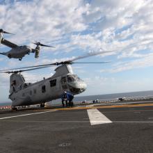 A helicopter with two sets of propellors placed on the sides of the aircraft flies over a helicopter on a tarmac with propellors on top of the body.