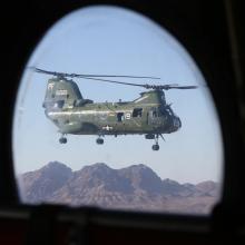 Porthole-style view of green military helicopter with two sets of propellors on top of the helicopter's body. The helicopter is in flight.