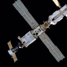 Bottom view of a section of the International Space Station with a cylinder-shaped module with one side attached to a smaller solar panel. The other side is attached to a much larger solar panel.