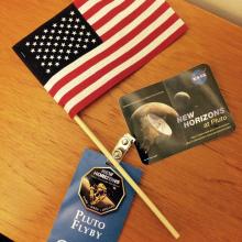 An American flag, blue guest badge, and sticker featuring Pluto that were given out at an event celebrating the New Horizons spacecraft.