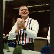 Eugene C. Kranz, a white male who served as Director of Missions Operations during Space Shuttle Mission STS-41C, wears a blue, red, and white vest in celebration. He is clapping.