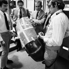 A group of five male NASA employees on Earth work together to come up with a solution for a wheel fender repair on the Lunar Module during the Apollo 17 mission.