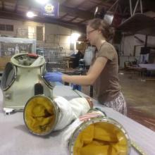 A white, female Museum intern works with spare spacesuit parts including arm sleeves and the neck piece.