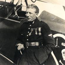 Charles Nungesser, a French World War I male pilot, poses in front of his aircraft and next to his famous insignia on the aircraft. The insignia consists of a skull, crossbones, coffin, and two candles inside a heart.