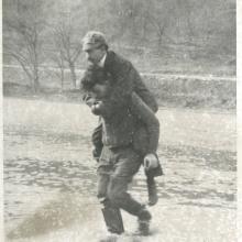 A mechanic working with Orville Wright gives Orville a piggyback as they walk in a river.