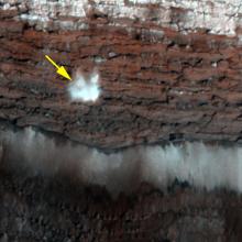 A frost-covered cliff on the northern polar surface of Mars. A yellow arrow points out potential frost that is in the process of freefalling.