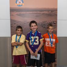 Three boys stand in front of a banner of Mars that is congratulating them on their completion of an astronaut game.