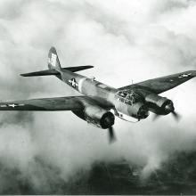 Diagonal front view of military monoplane with twin engines. U.S. star insignia is seen on the wings and rear fuselage. Aircraft is in flight.