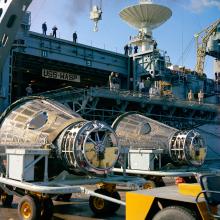 Frontal view of two conical-shaped spacecraft placed on their side as they are transported from a U.S. Navy carrier.