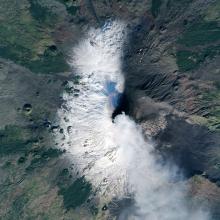 Satellite view of Mount Etna, a volcano in Italy, that is in the process of erupting. Ashes and gases are seen as a set of white clouds above the eruption site.