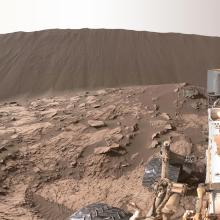 Tall sand dune in the background of a rocky, Mars surface. Curiosity Rover, a robotic surveyor, can be seen on the right side.