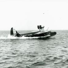 Side view of dark-colored sailplane with the name "U.S. Coast Guard" painted in a lighter color on the back fuselage of the aircraft. Two propellors can be seen on top of the front of the fuselage.