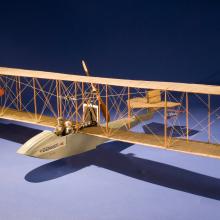 Wooden biplane-airboat model with space for two people, a passenger and the pilot. Flaglets are placed on the front structure in between the wings.