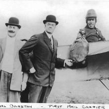 Earle Ovington, a male mail pilot, sits in the cockpit of an aircraft holding a bag of mail.