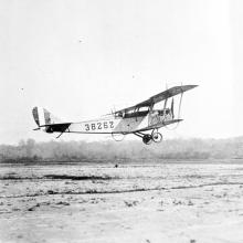 Side view of light-colored biplane in flight. Registration number "38262" written on back half of fuselage.