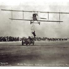 Front view of a biplane during an expedition flight. A person is trying to climb from an automobile below the biplane onto the biplane.