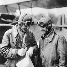 Two male military pilots in aviation gear look at a watch on the hand of the man on the left.