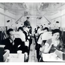 Interior of an early commercial aircraft. People sit in seats that have two rows of one seat apiece. The people in the front seem to have received some form of food service.