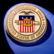 Golden-colored seal of the Civil Aeronautics Board featuring a red, white, and blue flag in the center below a bald eagle. The flag is covered in part by a yellow biplane.