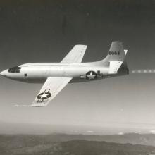 Side view of light-colored aircraft in flight. Aircraft features multiple U.S. military star inside circle with bar emblems.