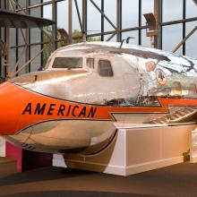 Nose, cockpit, and front fuselage section of a metal commercial airliner with orange, black, and silver-colored American Airlines livery.