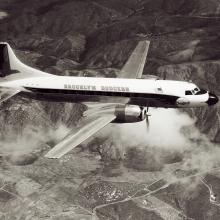 Side view of commercial aircraft in flight. Aircraft has two engines and features "Brooklyn Dodgers" name on the center of the side of the fuselage.