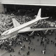 Top view of commercial jet with four engines and Boeing livery. People are crowded around the prototype jet.