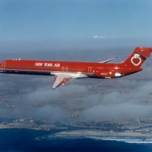 Side view of commercial jet in flight. Aircraft features red and white New York Air livery and two jet engines near the back of the plane.