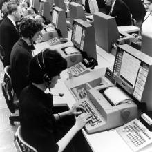 A group of people in an office setting use computers for airline reservations.