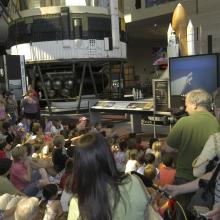 Museum visitors gathered around a large TV with Space Shuttle launch visible on the screen.