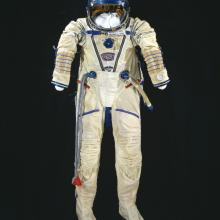 White pressure suit with blue trim and multiple plugs and tubes