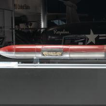 A long and skinny cylinder with a pointed tip at one end and tail fins at the other end. One half is painted red and the other half is left unpainted, showing the silver metal material it is constructed of.