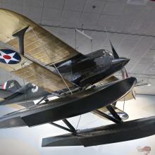 Black seaplane with pontoon floats instead of wheels and cream colored wings, hanging in museum