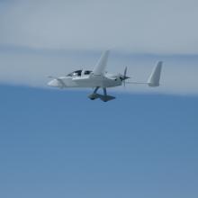 Side view of a white aircraft in flight with fixed landing gear and a propellor located on the rear of the fuselage.