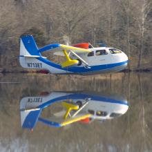 Side view of blue and white amphibious aircraft flying slightly above body of water. Registration number N713ET is painted in blue on the lower part of the vertical stabilizer.